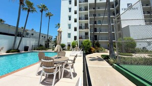 A pool area with lounge chairs and umbrellas, a fenced tennis court, and a multi-story building with balconies. - Beachfront 2BR, 5th Floor Views, Wakable SPI (South Padre Island)