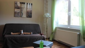 Ein Wohnzimmer mit einer Couch, einem kleinen Tisch mit einer grünen Vase, einer Stehlampe und einem Fenster mit Vorhängen.