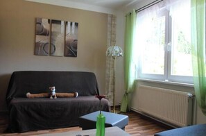 Ein Wohnzimmer mit einer Couch, einem kleinen Tisch mit einer grünen Vase, einer Stehlampe und einem Fenster mit Vorhängen.