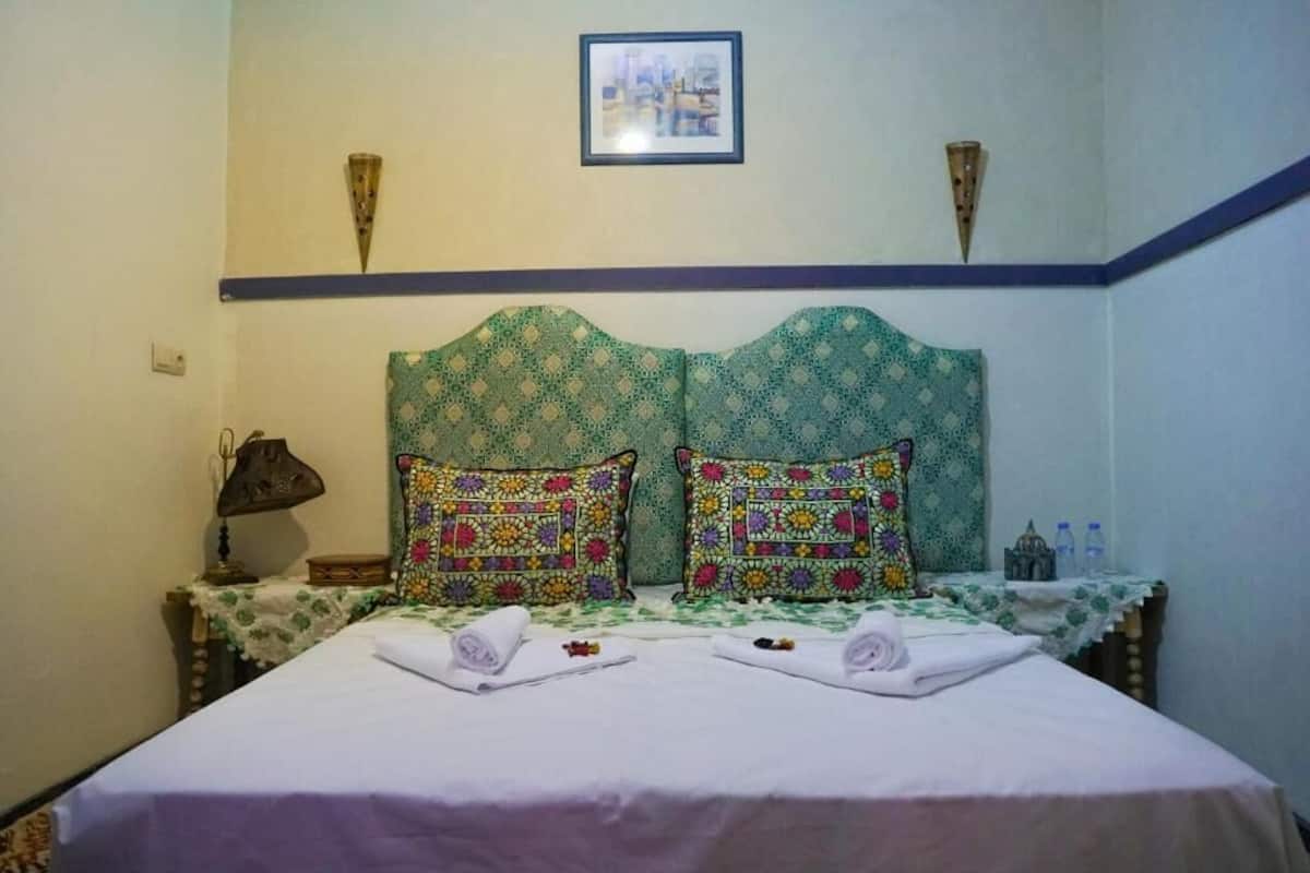 A bed with a patterned headboard, two pillows, a bedside table with a lamp, and a framed picture on the wall.