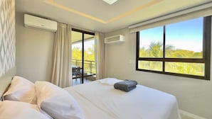 Una habitación de hotel con cama, ventana con vista a palmeras, unidad de aire acondicionado y una lámpara de techo.