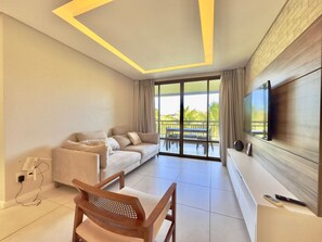 Ein modernes Wohnzimmer mit einer Couch, einem Holzstuhl, einem Flachbildfernseher und einem Balkon mit Gartenmöbeln.