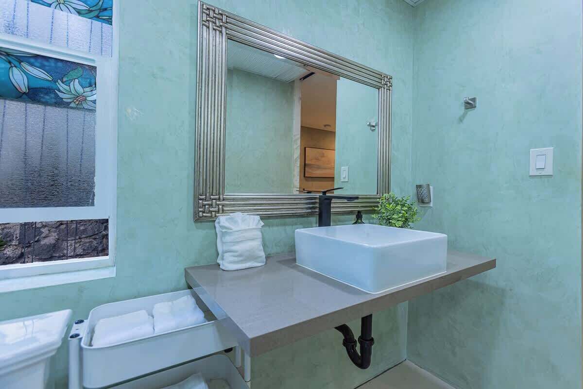 Une salle de bain avec un lavabo blanc, un miroir doté d’un cadre décoratif et une fenêtre donnant sur un mur en pierre.