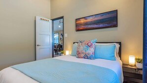 A bedroom with a bed, a painting, a door, and a nightstand with a lamp. - Charming 2BR Condo in Historic Wailuku | Central Maui Location (Wailuku)