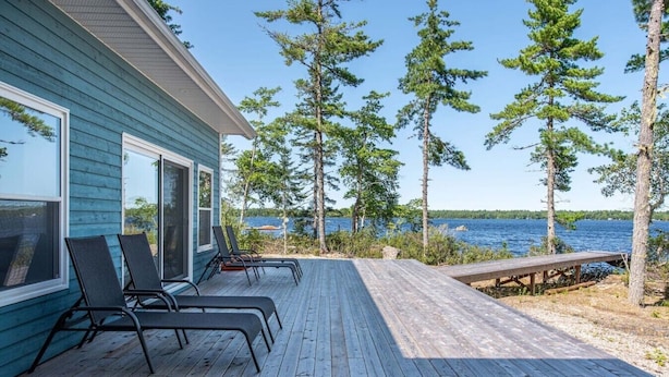 A wooden deck with lounge chairs, a wooden bench, and a house with large windows overlooking a body of water.
