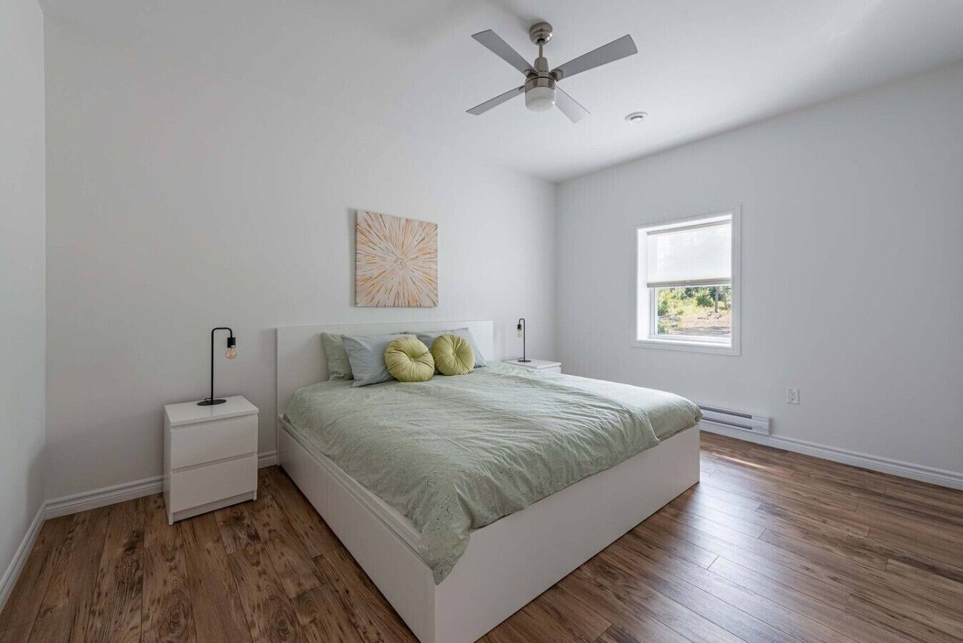 A bedroom with a bed, nightstands, a ceiling fan, a window, and a wall-mounted artwork.