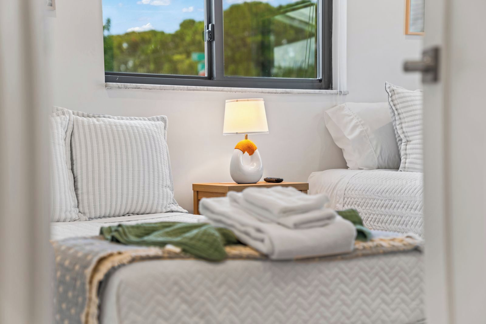 A bedroom with a bed, pillows, a bedside table with a lamp, and a window with a view of greenery.