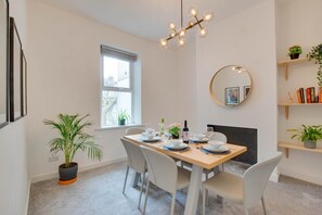 A dining area with a table set for four, a potted plant, a window with blinds, a wall-mounted mirror, and a bookshelf with books and plants.