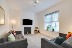 A modern living room with two grey sofas, a flat-screen TV mounted on the wall, a fireplace, and a large window with blinds.