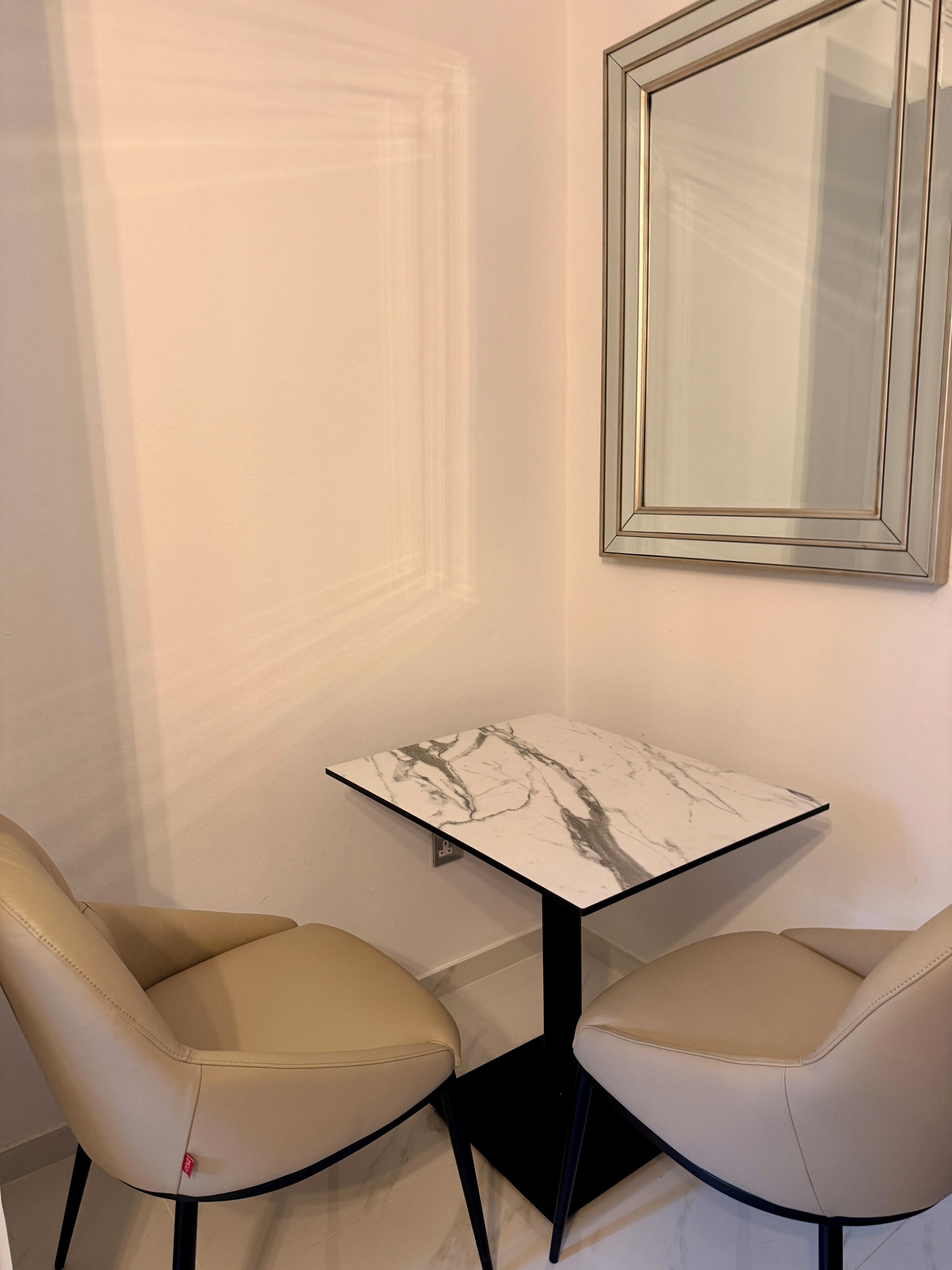 A small, square table with a marble-like surface, flanked by two beige chairs with black legs. A framed mirror hangs on the wall above the table.