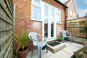 A brick house with a white door, two grey chairs, a potted plant, and a wooden fence. - 1 Bed in Burnham Market (oc-2193) (Burnham Market)