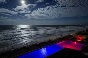 A nighttime view of a beach with a pool illuminated by red and blue lights, a fence, and a distant moon. - Opulent Ocean view with heated pool and hot tub (Daytona Beach)