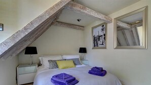 Een slaapkamer met een schuine kap, een bed met kussens, twee nachtkastjes met lampen, en een ingelijst schilderij aan de muur.