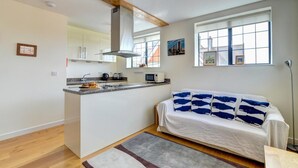 A compact living space with a kitchenette, a sofa with patterned cushions, and a view of the city through large windows. - 2 Bed in Swanage (oc-wy412) (Swanage)