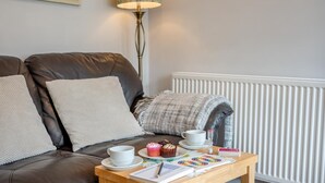Un salon chaleureux avec un canapé en cuir marron, un radiateur blanc, une petite table en bois sur laquelle se trouvent une tasse, un cupcake et un jeu de société.