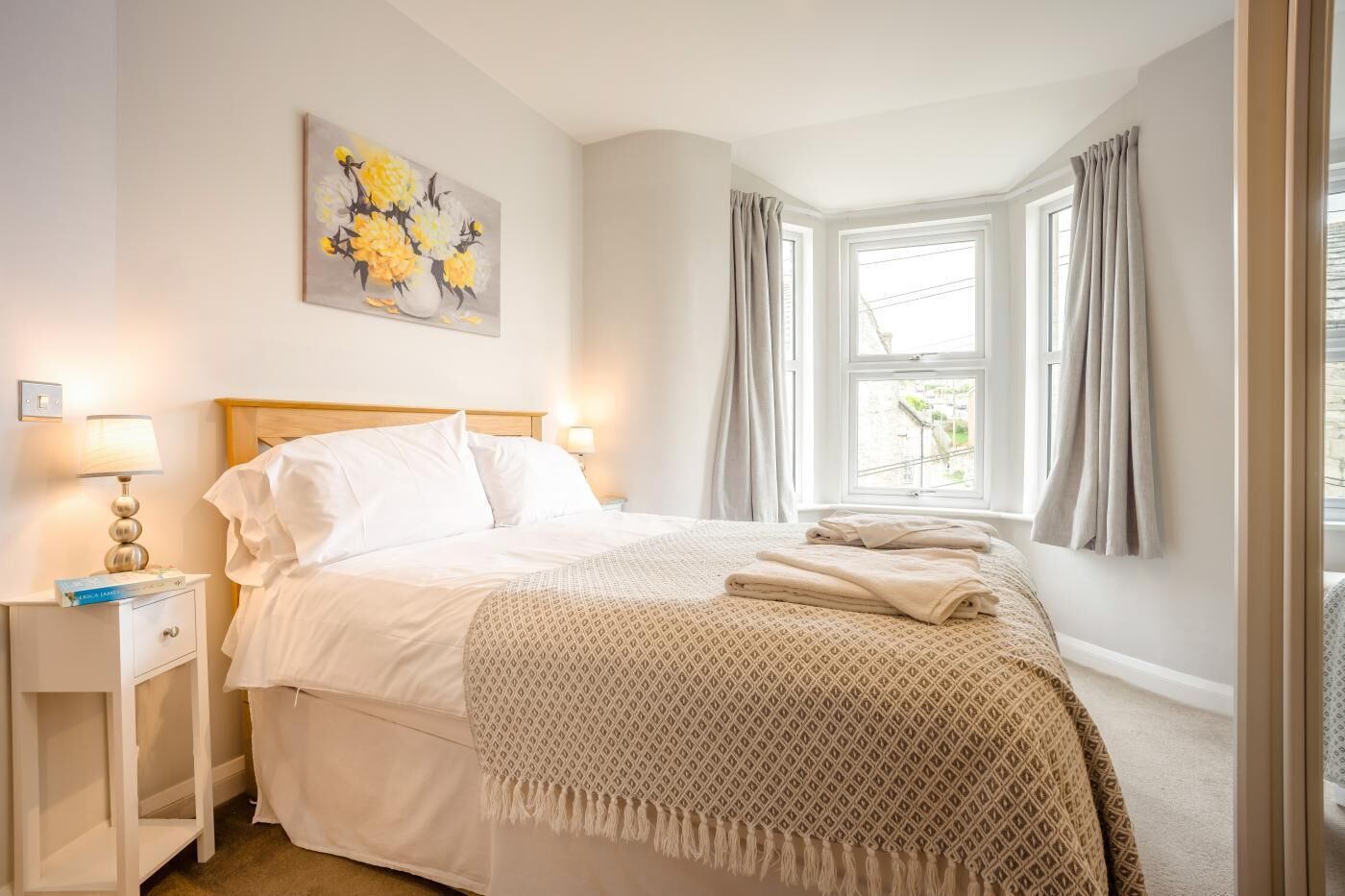 A neatly made bed with a patterned blanket, a bedside lamp, a nightstand with a book, and a painting of flowers on the wall.