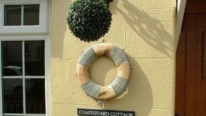Una cabaña costera con un flotador salvavidas y un letrero que dice 'COASTGUARD COTTAGE' en la pared.