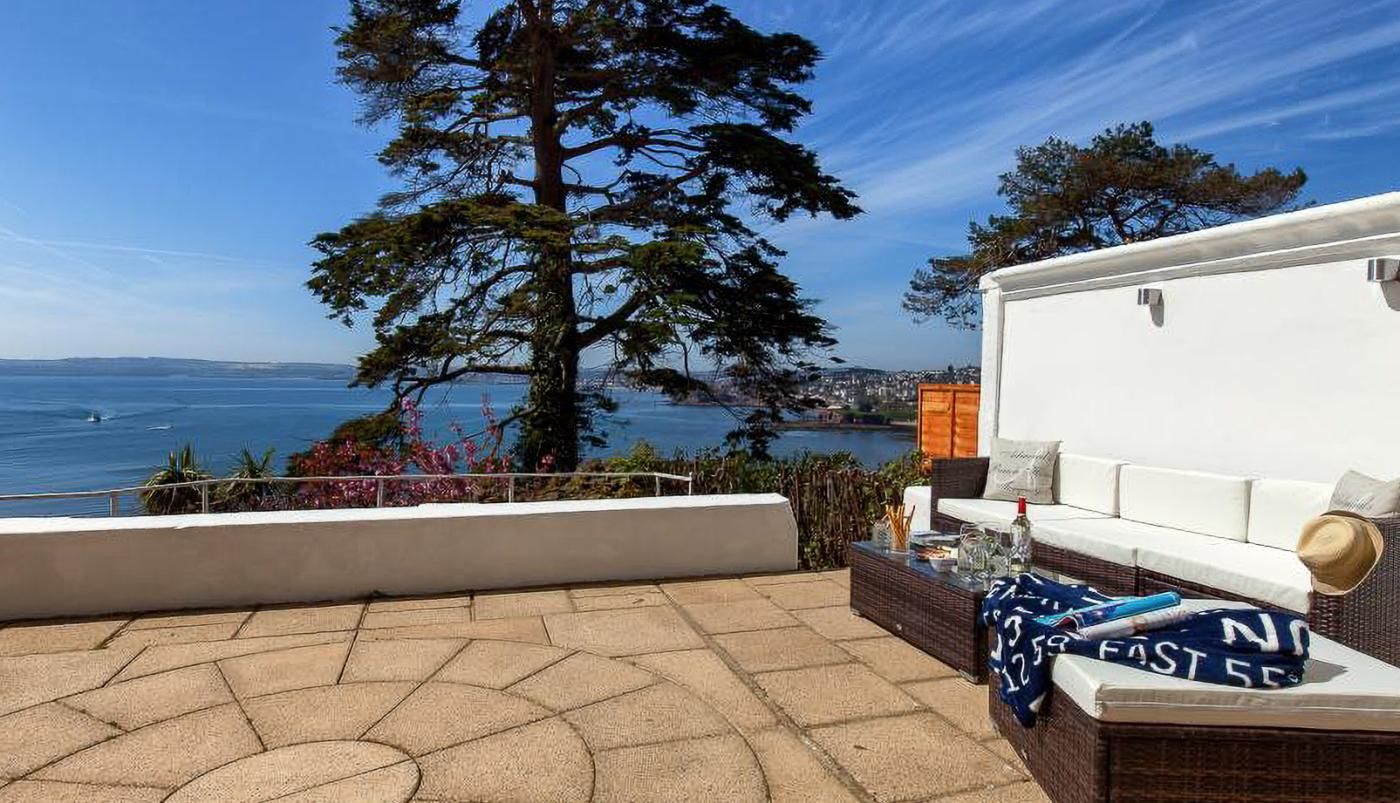 A patio with a view of the ocean, a white sofa, and a wicker table with a blanket.