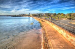 Eine Uferpromenade mit einer Steinmauer, einer parallel zur Wasserlinie verlaufenden Straße und einer Ferne mit Gebäuden und Bäumen.