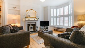 A living room with a fireplace, sofas, a television, and a large window with shutters. - 3 Bed in Mumbles (oc-hh1223) (The Mumbles)