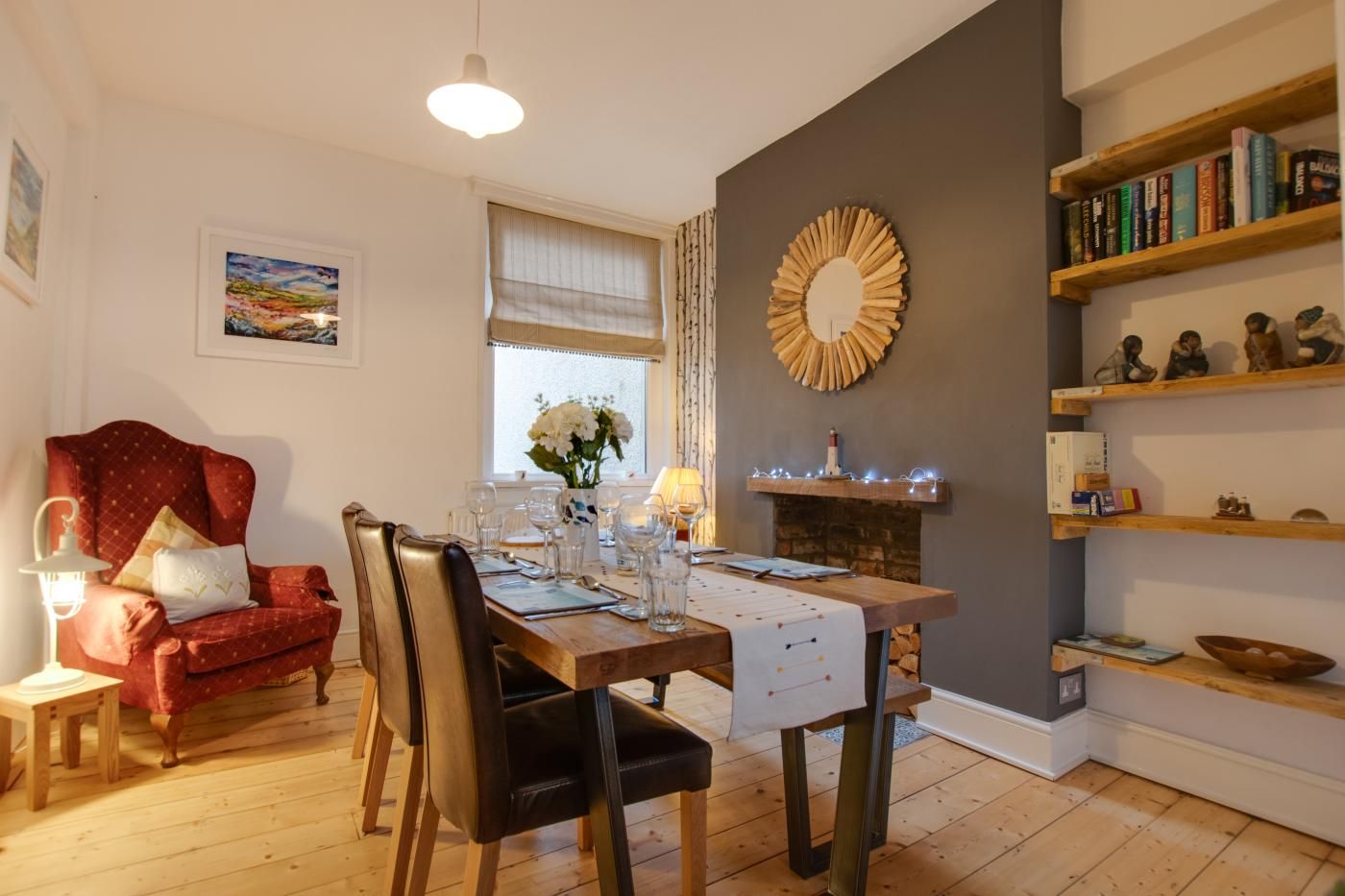 A dining area with a wooden table set for four, a red armchair, and a fireplace with a decorative mirror.