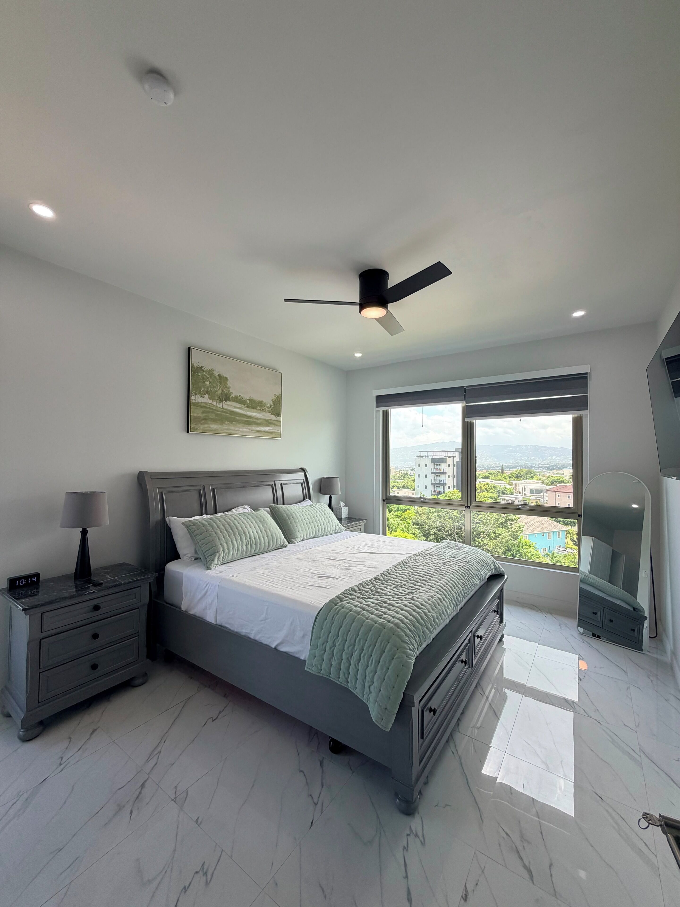 A modern bedroom with a bed, nightstand, lamp, ceiling fan, and a large window offering a city view.