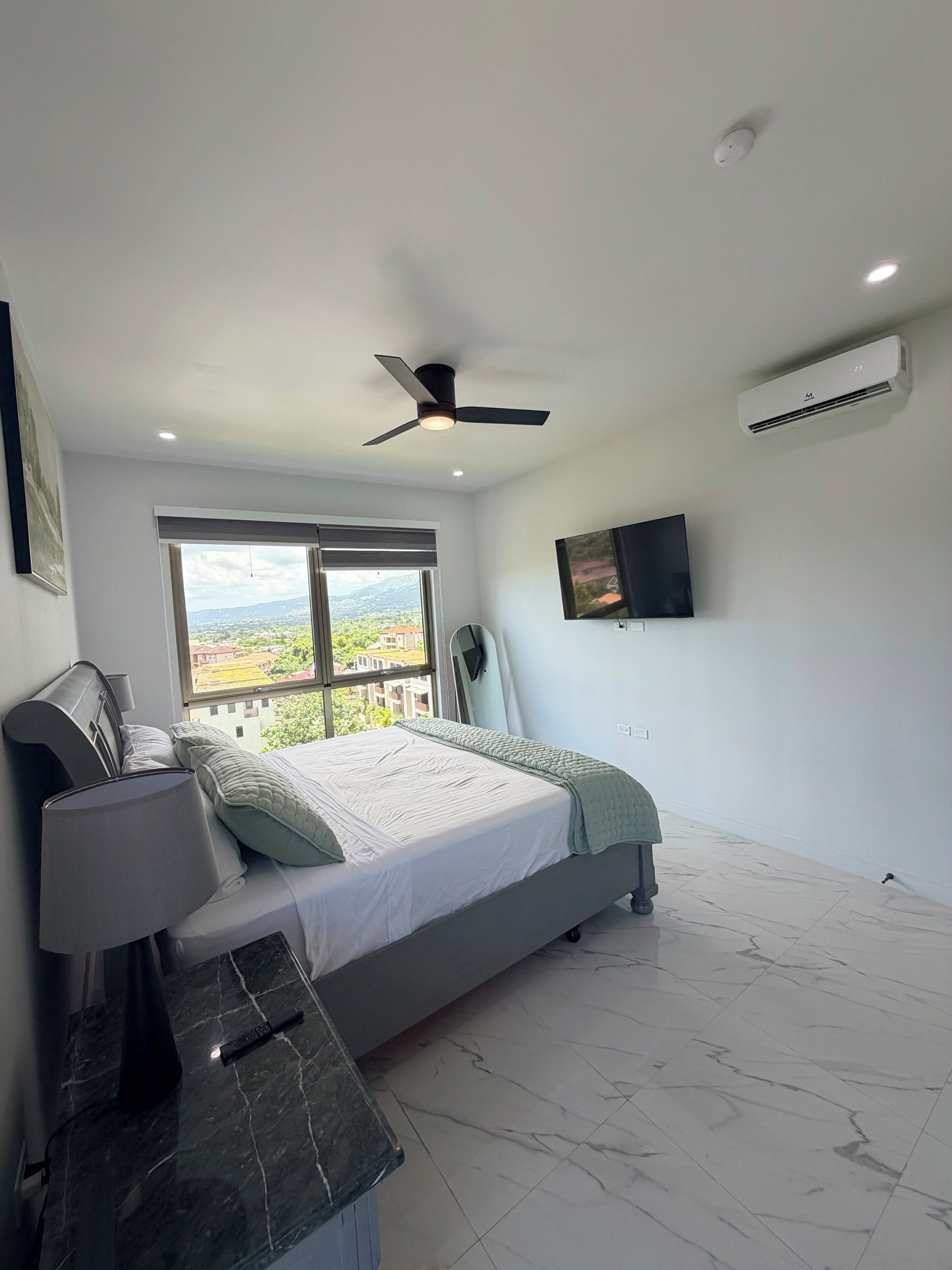 A bedroom with a bed, a ceiling fan, an air conditioning unit, a TV, and a window with a view of buildings and greenery.