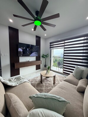 A modern living room with a ceiling fan, a flat-screen TV mounted on the wall, a sofa, a coffee table, and a large window with blinds. - 2BDRM • 24hrs Security • Top floor with Mountain View • Rooftop Pool and Gym (Kingston)