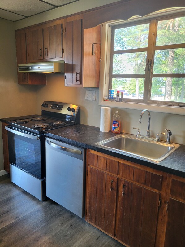 A kitchen with wooden cabinets, a stainless steel stove, a dishwasher, a sink, and a window with a view of trees.