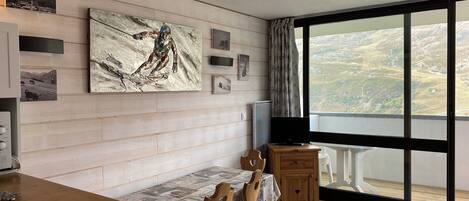 Een eethoek met een houten tafel en stoelen, een televisie en een groot raam met uitzicht op een glooiend landschap.