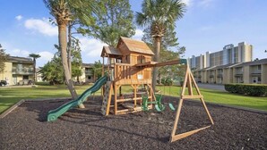 A playground with a wooden slide and swings, surrounded by palm trees and residential buildings. - 4 bed 2 bath townhome walking distance from the beach! (Panama City Beach)