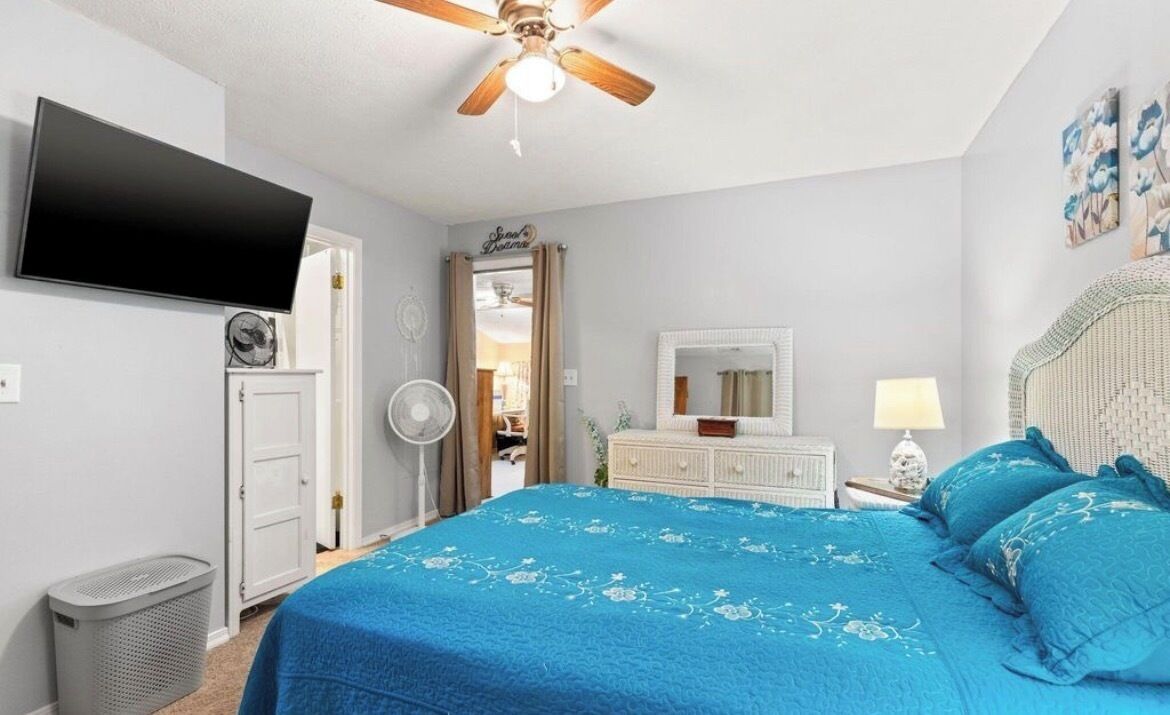 A bedroom with a bed, a ceiling fan, a TV, a dresser, a mirror, and a door leading to another room.