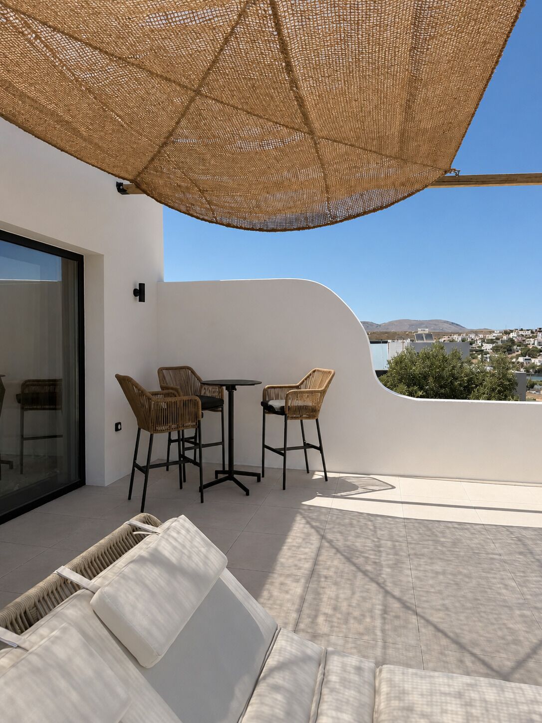Ein moderner Außenbereich mit Sitzgelegenheit, Tisch und Stühlen, einem großen gewebten Sonnenschirm und Blick auf die Stadt.