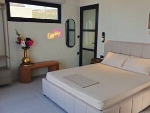 Une chambre à coucher avec un lit, une table de chevet ornée d’un vase de fleurs, un banc, un panneau lumineux au néon et une fenêtre.