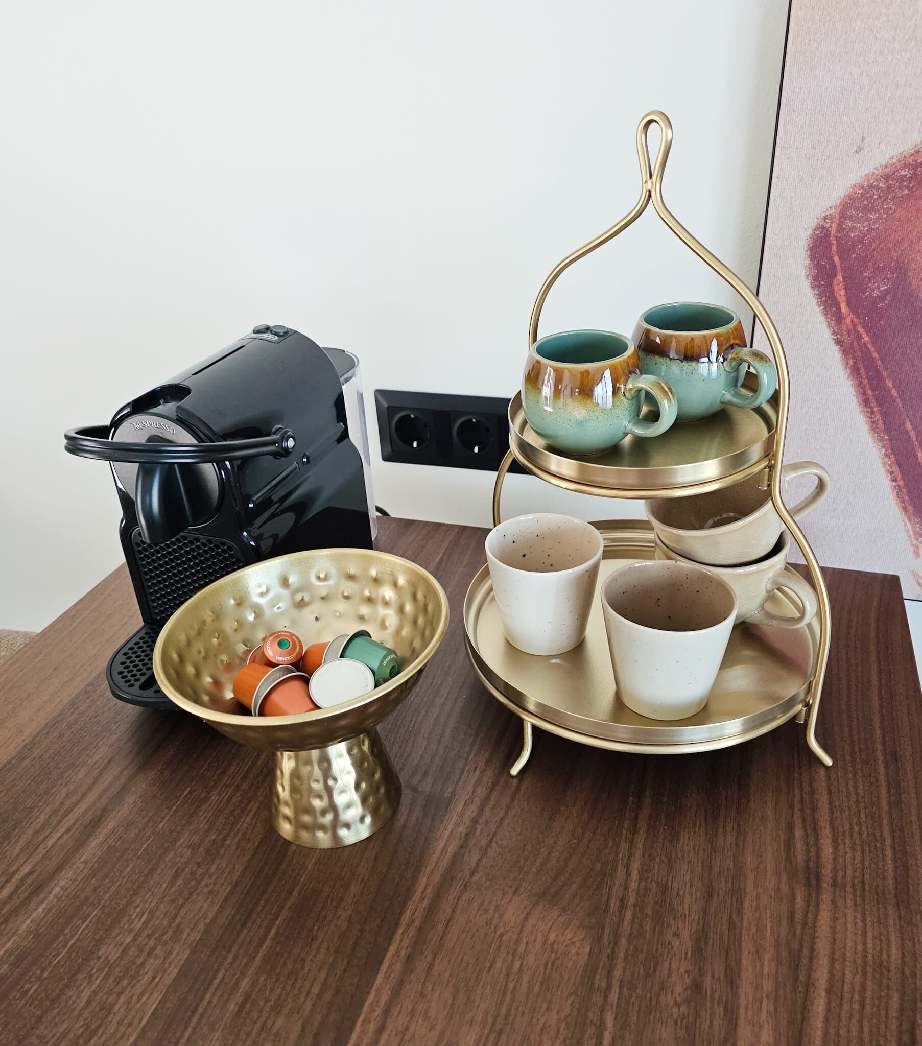 Una cocina con cafetera, un frutero de tres niveles con tazas y un bol de cápsulas de café.