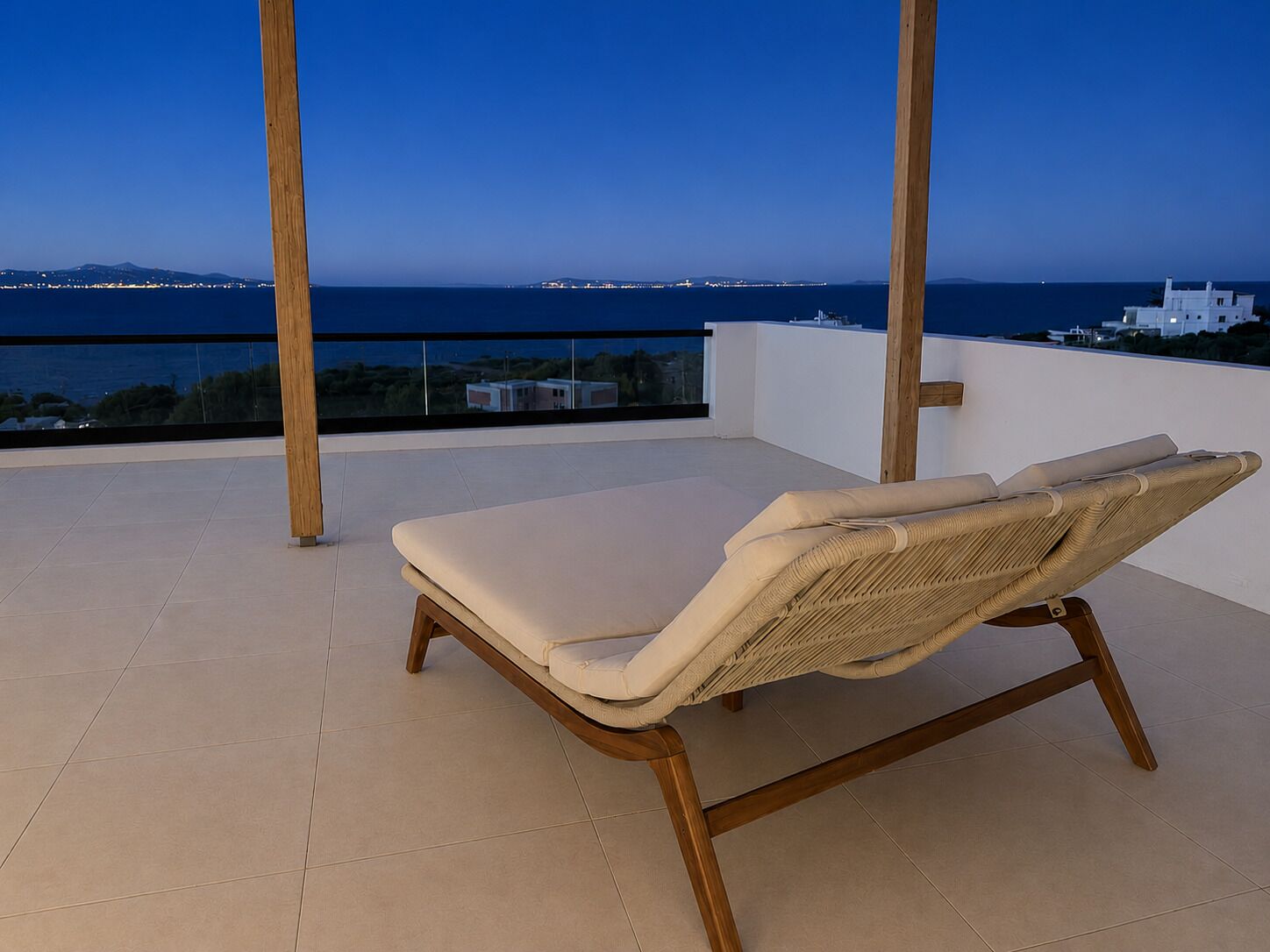 A lounge chair on a balcony with a view of the sea and distant city lights.