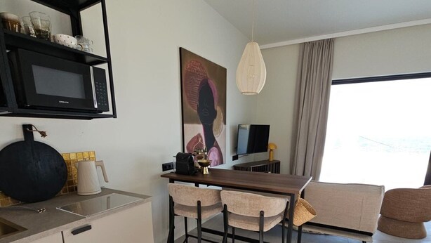 A modern kitchen with a microwave, dining table, and chairs. There is a painting on the wall, a television, and a window with curtains. - Syros House - Suite: Slow Living by the Sea, Private Terrace, Ocean Views (Azolimnos)