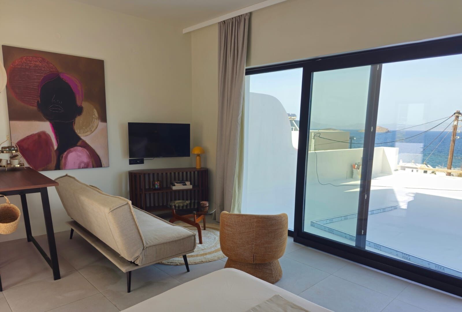 A modern living room with a sofa, a TV, a dining table, and a large window with a view of the sea.