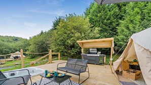 Ein überdachter Außenbereich mit Grill, Sitzgelegenheiten und Blick auf einen bewaldeten Hügel.