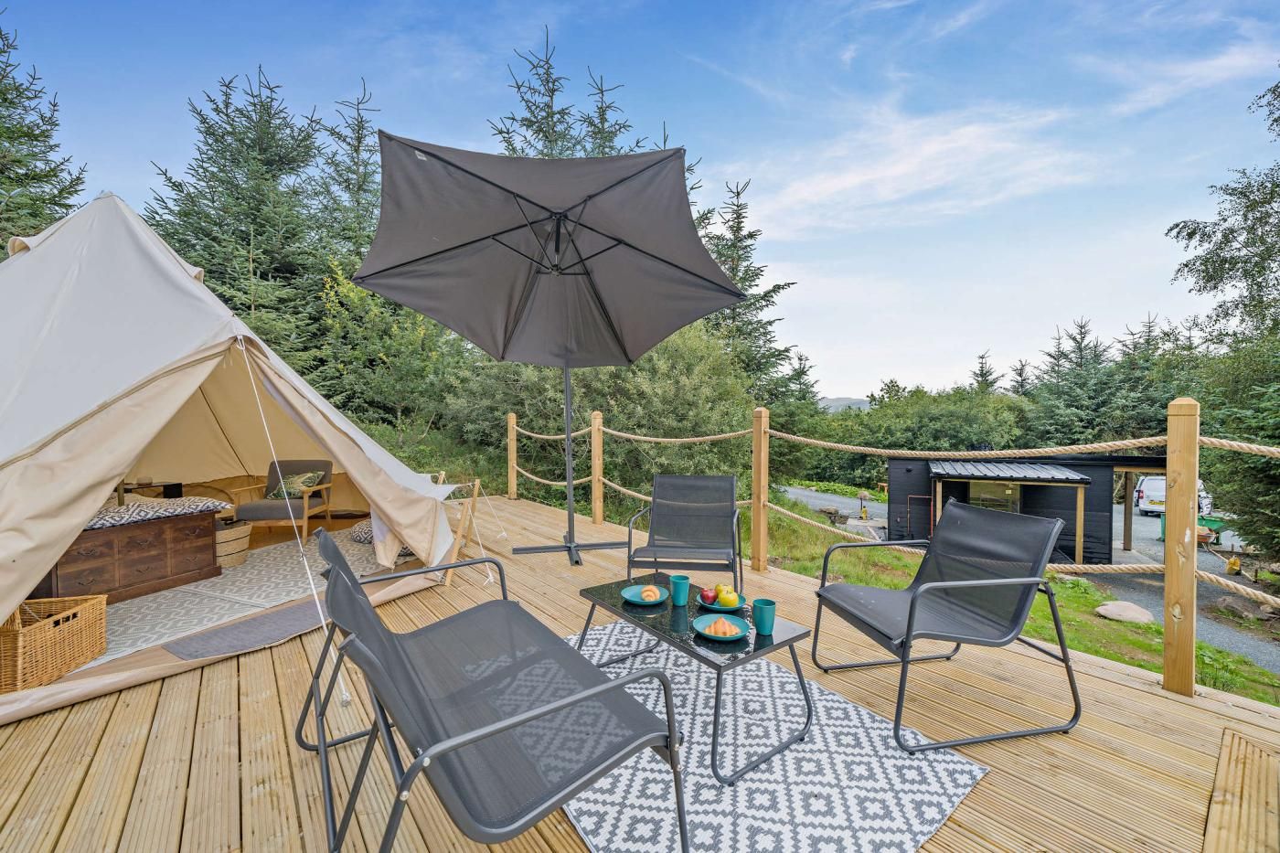 A tented accommodation with a deck, outdoor seating, and a small building in the background.