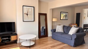 A living room with a flat-screen TV, a grey sofa, a white round table, and a framed abstract painting on the wall.