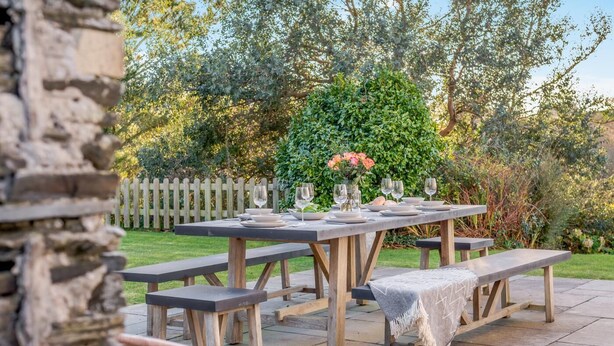 An outdoor dining area with a wooden table set for four, benches, and a stone wall in the foreground. - 4 Bed in Trethevy (oc-s33745) (Bossiney)