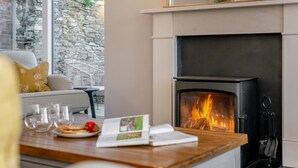 A cozy room with a fireplace, a dining table with glasses and a plate of food, and a sofa with cushions. - 4 Bed in Trethevy (oc-s33745) (Bossiney)