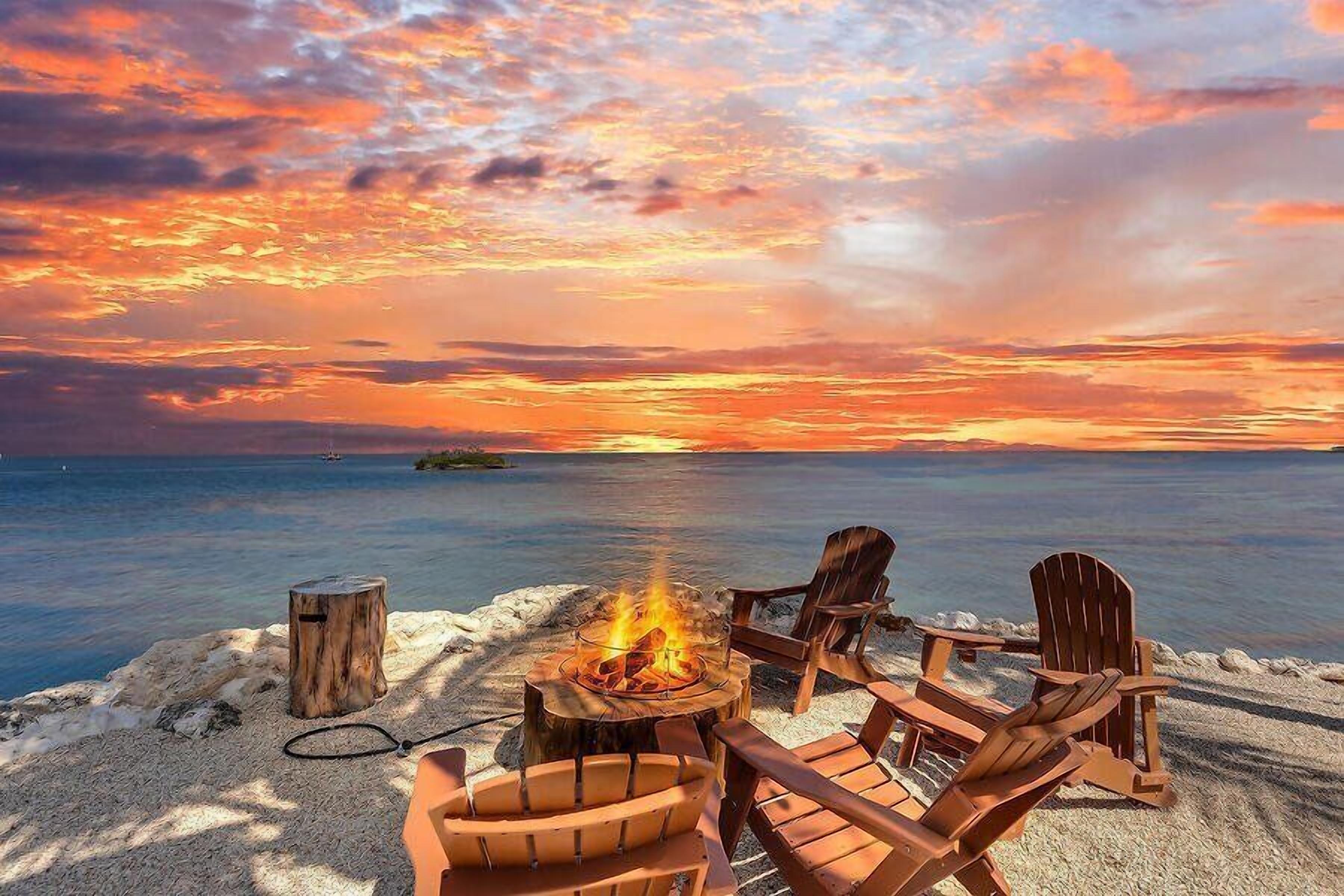 A beach setting with a fire pit and chairs facing the ocean, under a sunset sky.