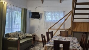 Una habitación con sofá, mesa de comedor, sillas, televisor montado en la pared, escalera y una ventana con cortinas.