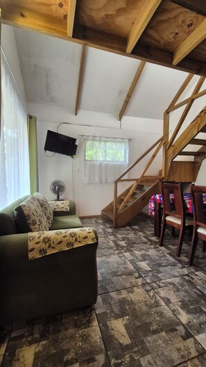 Una habitación con una escalera de madera, un sofá con cojines estampados, un ventilador y una ventana con cortinas.