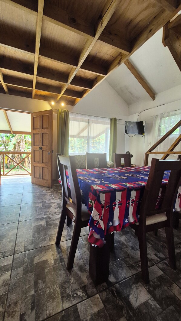A room with a wooden ceiling, a bed with a colorful quilt, a TV mounted on the wall, and a view of the outdoors through a window.