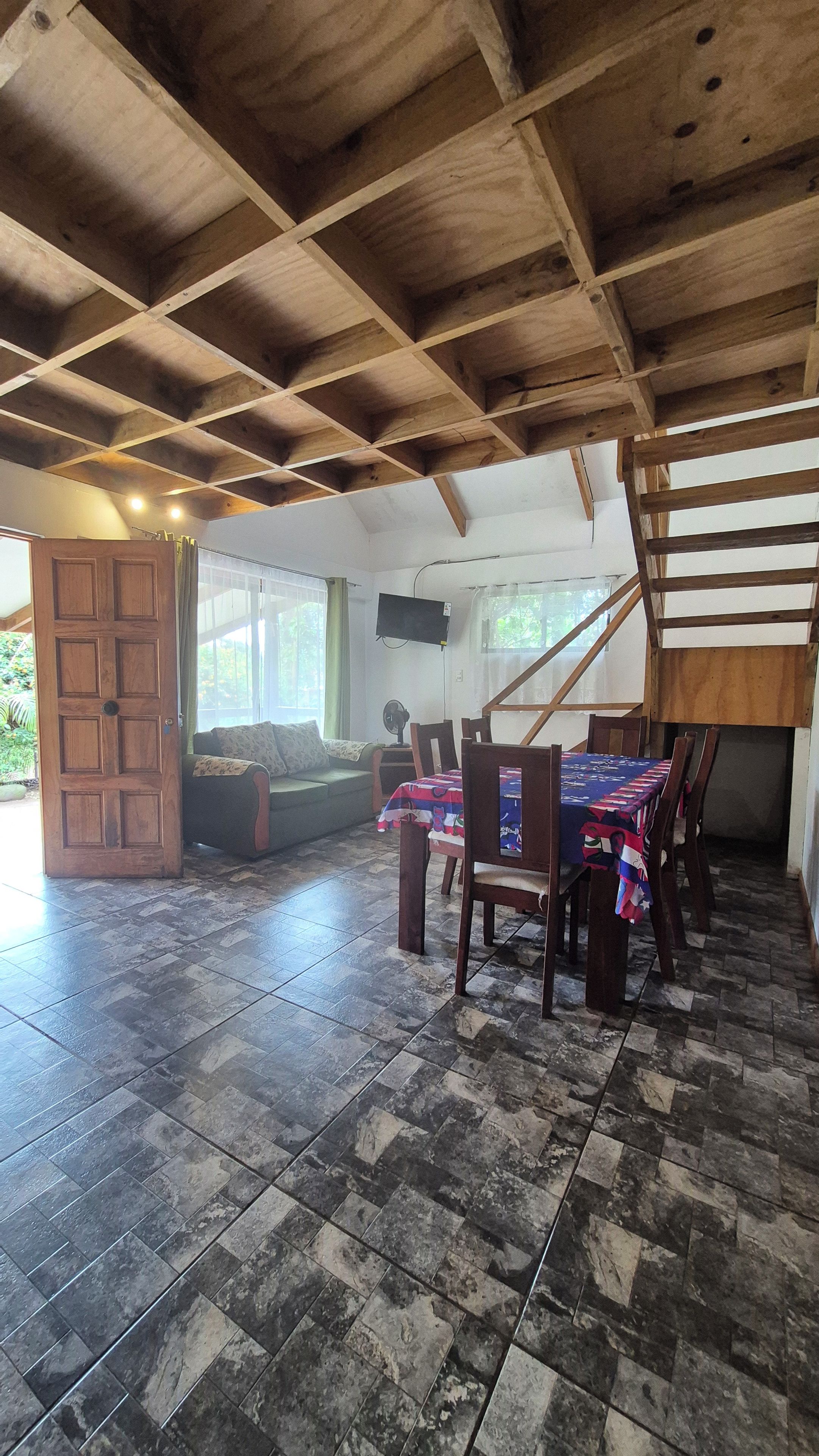 A spacious room with a high ceiling, a dining table with chairs, a sofa, and a television mounted on the wall.