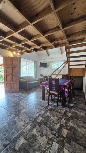 A spacious room with a high ceiling, a dining table with chairs, a sofa, and a television mounted on the wall.