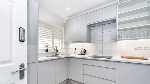 Eine moderne Küche mit weißen Schränken, einer Edelstahlspüle, einem Wasserkocher und einem Fenster mit Jalousien.